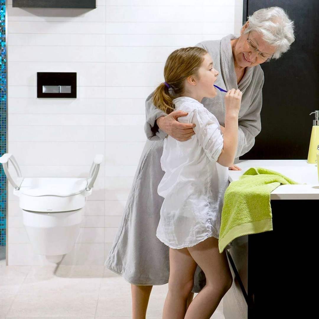 Etac Cloo adjustable height toilet seat raiser mounted on toilet with elderly woman hugging a young girl brushing teeth